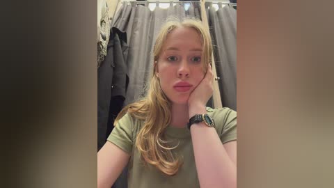 A video of a young woman with long blonde hair, wearing an olive-green t-shirt, leaning on her hand in a bathroom with grey towels hanging in the background.