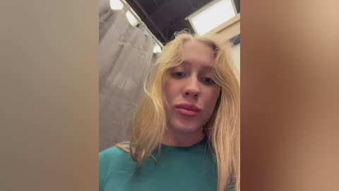 A video of a blonde woman with fair skin and a round face, wearing a teal shirt, in a dimly lit bathroom with beige walls and a shower curtain.