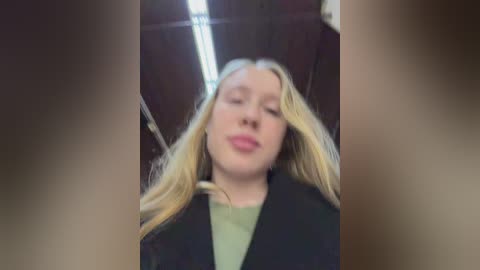 A video of a fair-skinned, blonde woman with long hair, wearing a green top and black jacket, smiling with her eyes closed, taken indoors with a blurred background.
