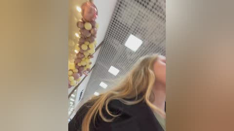 A blurry video of a blonde woman with shoulder-length hair, wearing a black jacket, looking away from the camera in a brightly lit, modern hallway with a decorative yellow and brown balloon garland on the ceiling.