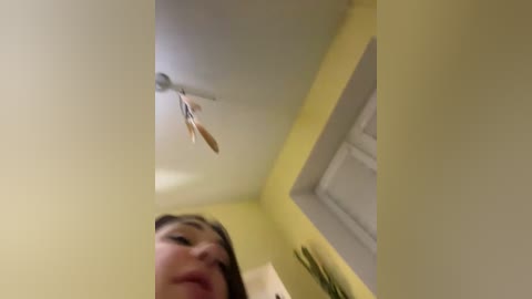 A video capturing a close-up view of a woman with long, dark hair, standing in a bright yellow-painted room with a ceiling fan and a white door.
