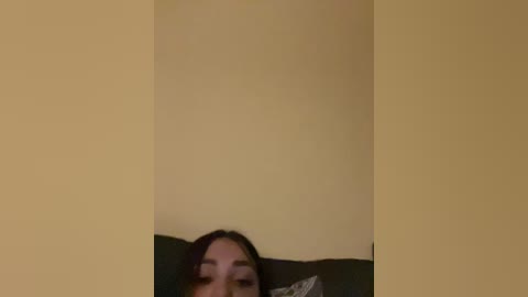 A video of a woman with dark hair and fair skin, partially visible, lying on a dark couch against a beige wall. The image is slightly blurred.