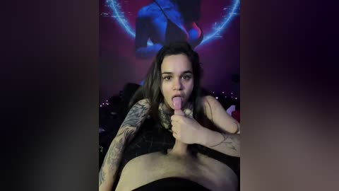 Video of a tattooed woman with long dark hair licking a man's erect penis in a dimly lit room with a neon sign in the background.