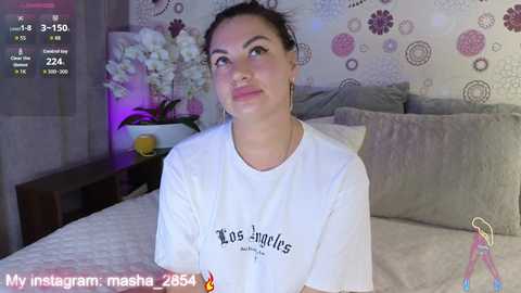 A video of a young woman with light skin and dark hair, wearing a white t-shirt, sitting on a bed in a room with floral wallpaper.