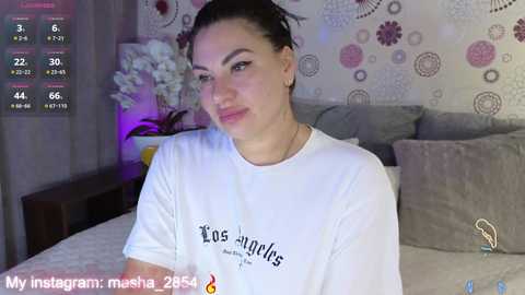 A video of a young woman with dark hair tied back, wearing a white t-shirt with \"Los Angeles\" text, in a cozy bedroom with floral wallpaper, grey pillows, and a nightstand.