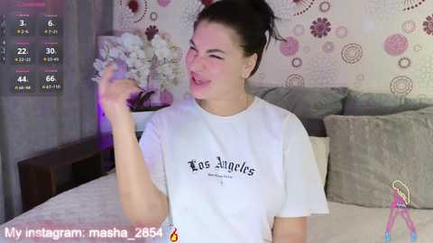 Video of a woman with dark hair, wearing a white t-shirt, smiling in a bedroom with floral wallpaper, bed, and pink lighting.