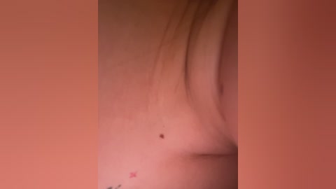 Video of a close-up of a person's upper chest and shoulder with a mole near the left nipple. The skin is fair with a few minor blemishes. The background is a muted, warm tone, possibly peach or coral.