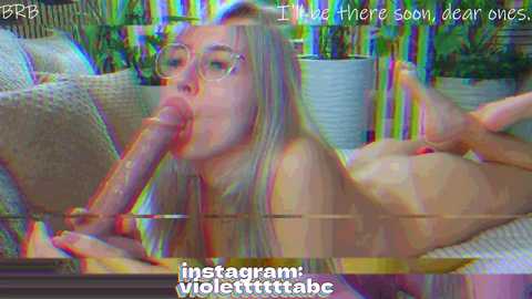 A video of a nude woman with glasses and long blonde hair performing oral sex on a large dildo, set in a modern living room with plants.