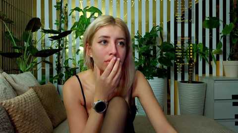 Video of a young blonde woman with fair skin, wearing a black tank top and a smartwatch, covering her mouth in surprise. Green plants, white furniture, and a striped background create a cozy, modern interior.