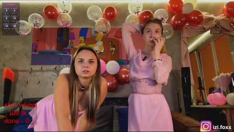 Video of two young girls in pink dresses, balloons, and a messy room. One girl, with long hair, leans forward, looking worried.