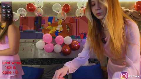 Video of a young woman with long blonde hair in a pink dress, helping a girl with long brown hair in a pink dress. Background shows a colorful party scene with balloons and decorations.