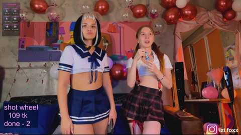 Video of two young women in a cozy room, one in a sailor schoolgirl outfit, the other in a plaid skirt. Decorated with balloons, colorful walls, and a TV in the background.