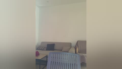Video of a minimalist living room with beige couches, a light blue plastic chair, and a small wooden coffee table. The room has neutral walls and a single, small pink object on the floor.
