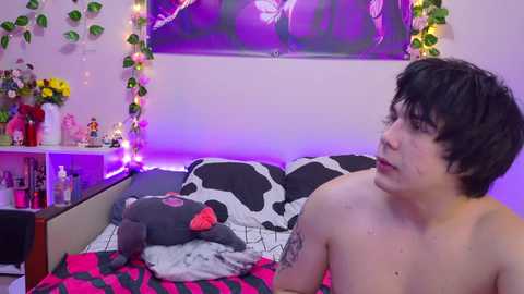 A video of a shirtless, pale-skinned person with short, dark hair and a tattoo on their left arm, sitting on a bed with a cow-patterned duvet, surrounded by fairy lights, a stuffed animal, and a framed poster in a cozy bedroom.