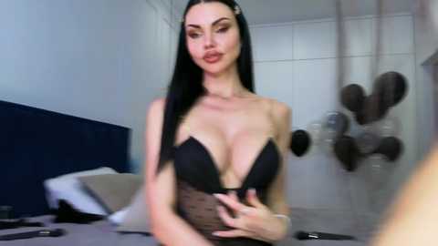 Video of a slender, pale-skinned woman with long black hair, wearing a black strapless bra, standing in a modern, white-tiled bathroom. The background shows a bed with pillows, and a mirror reflecting her image.