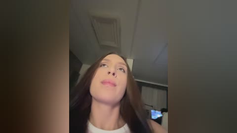 A video of a young Caucasian woman with long brown hair, wearing a white tank top, looking upward in a dimly lit room with a white ceiling and a window in the background.