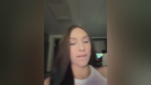 A blurry video of a young woman with long brown hair, fair skin, and a white tank top, standing in a dimly lit room with a TV and a couch in the background.