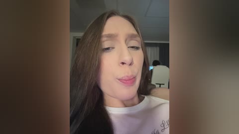 Video of a young, light-skinned woman with straight, long brown hair, wearing a white T-shirt, making a playful pouty face in front of a blurred, dimly lit room with a chair and a door.