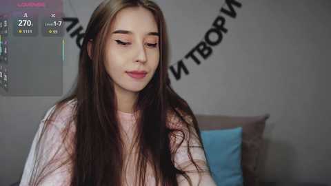 Video of a young Caucasian woman with long, straight brown hair, wearing a light pink sweater, smiling softly, in a cozy bedroom with a gray wall and a \"Happy Birthday\" banner.