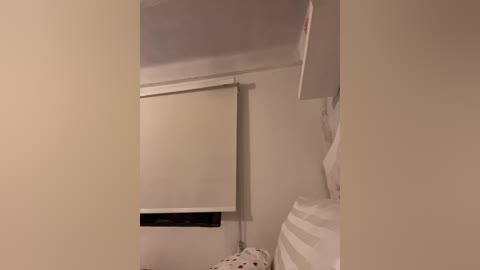 A video of a beige wall with a white roller shade partially covering a window. A white pillow with red polka dots is visible on the floor.
