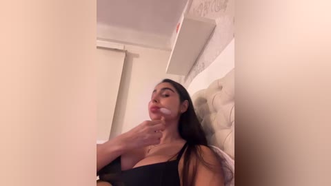 A video captures a Latina woman with long black hair, wearing a black bra, sitting on a beige bed. She has a light tan skin tone, and her face is covered in whipped cream, giving a playful, sensual vibe.