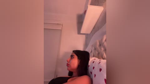 A video of a Latina woman with medium brown skin, long black hair, and a black tank top, lying on a bed with a heart-patterned pillow, surrounded by white walls and a tufted headboard.