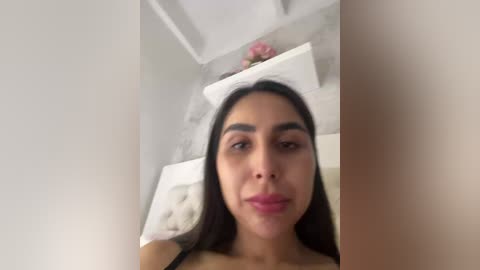 Video of a Latina woman with long, straight black hair, light brown skin, and full lips, wearing a black top. She stands in a modern, minimalist room with white furniture and a vase with pink flowers on a shelf.