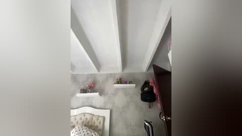 Video of a minimalist, modern bedroom with white walls, high ceilings, and exposed wooden beams. The room features a tufted beige headboard, a small white shelf with a pink vase, and a black backpack. The floor is concrete.