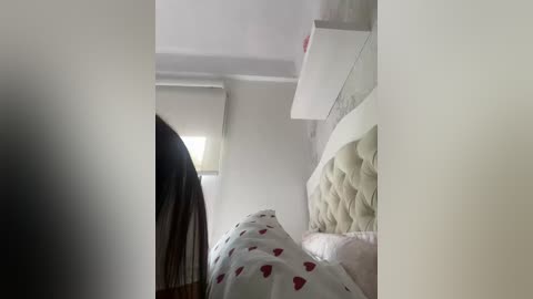 A video of a minimalist bedroom with a beige tufted headboard, white bedding with red heart pattern, and a white floating shelf.