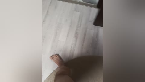 A video of a nude person with light skin tone, standing in a corner, looking down at a beige carpeted floor. The person's feet are visible, and the background includes a white wall and a wooden door.