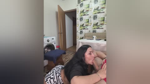 Video of a woman with long black hair performing oral sex on a man in a small, cluttered bedroom with a white washing machine, a bed, and a wall with a newspaper collage.