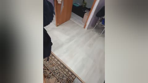 A video shows a person in dark pants and a white shirt standing on a white-painted floor, with a patterned rug and wooden furniture in the background.