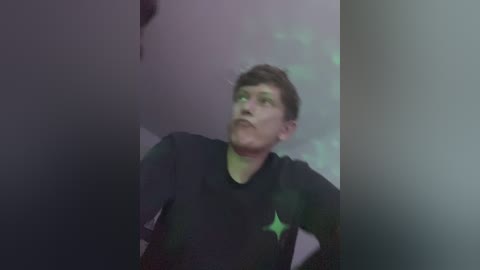 A video of a man with short brown hair, wearing a black shirt, looking upwards in a dimly lit room with blurred green and purple hues.