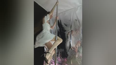 Video of a young woman in a white dress and black high heels performing a pole dance, surrounded by reflective surfaces and a mirrored ceiling, creating a distorted, surreal ambiance.
