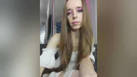 A video of a young, slender woman with long, straight brown hair, wearing a white off-shoulder dress and vibrant purple eyeshadow, seated in a dimly lit room with reflective surfaces.