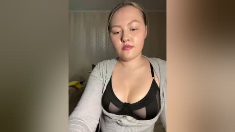 A video of a fair-skinned woman with light brown hair, wearing a black lace bra and a gray cardigan, sitting indoors, looking down.