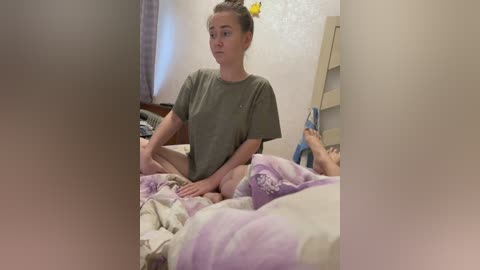 Video of a young woman with light skin, brown hair in a bun, wearing a loose green t-shirt, sitting cross-legged on a bed with pastel-colored sheets, in a dimly lit, cluttered bedroom.