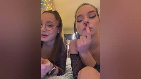 Video of two young women with fair skin, one with glasses, wearing sparkly black tops, kissing in a dimly lit room with a festive, colorful backdrop.