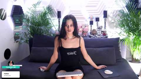 Video of a young woman with long black hair, wearing a black dress and sitting on a black couch in a modern, brightly-lit room with green plants and candles in the background.