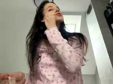 Video of a woman with long dark hair, fair skin, wearing a pink floral pajama top, adjusting her hair in a bathroom with white tiles and a skylight.