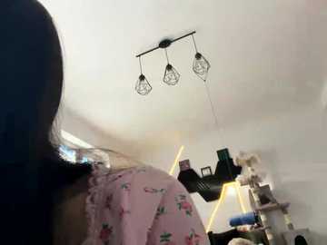 Video of a woman with long hair in pink pajamas, facing away, in a dimly lit room with white walls, a hanging light fixture, and a cluttered bookshelf.