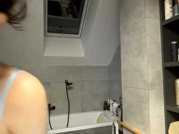 Video of a modern bathroom with a person's back, wearing a light blue tank top, standing near a white bathtub. Gray tiled walls and a skylight above. Shelves hold toiletries and a wicker chair.