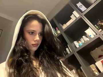 A video of a young woman with long, wavy black hair, wearing a white robe, standing in front of a dark grey shelving unit filled with cosmetics.