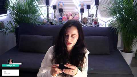 A video of a woman with long black hair, wearing a white hoodie, intently using a smartphone on a dark gray couch, surrounded by green plants and a display of plush toys in the background.