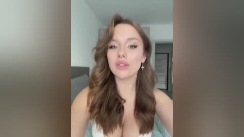 Video of a Caucasian woman with fair skin and wavy brown hair, wearing a low-cut white top, standing in a modern, minimalist room with a gray couch and white walls.