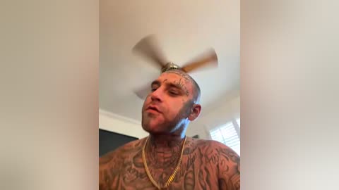 Video of a bald, muscular, tatted man in a gold chain, spinning a ceiling fan with his head, in a brightly lit room.