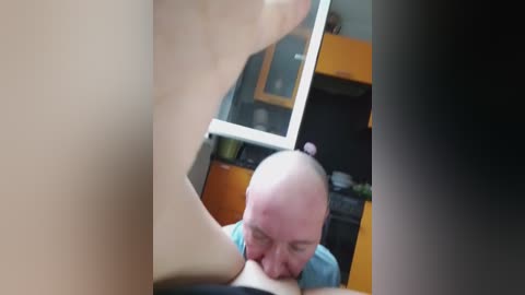 A video depicts a bald, middle-aged man with a light skin tone performing oral sex on a woman with a visible, prominent breast. The background shows a modern kitchen with wooden cabinets and a glass door.