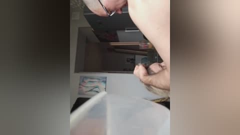 A video of a man with glasses, holding a woman's penis, in a messy room with artwork and a closet.