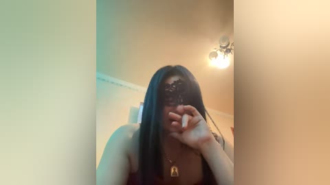 A video of a young woman with long black hair, wearing a black lace mask, posing indoors with a blurred background. She is partially dressed, with a visible necklace, and her right hand covers her face.