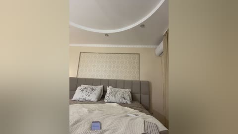 Video of a minimalist, beige-tiled bedroom with a modern, upholstered gray headboard. The room features a white ceiling fan, air conditioning unit, and patterned wallpaper. A smartphone lies on the bedspread.
